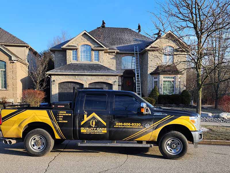Quality Care Roofing crew members working on the roof of a mid-class, two-story stone home with their work pickup parked in front